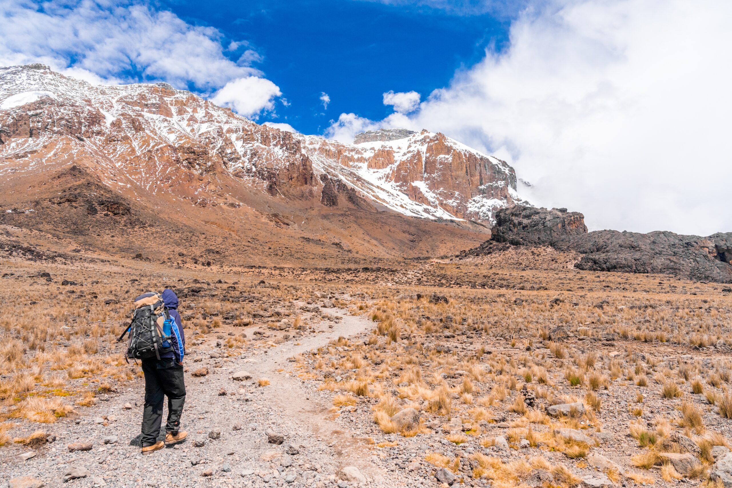 A horizontal shot of a hiker with backpack hiking in a dry field surrounded by rocks and mountains